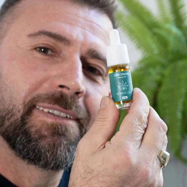 Mann hält Flasche Nordic Öl CBD Öl 5% mit Pipette in der Hand vor grünem Hintergrund